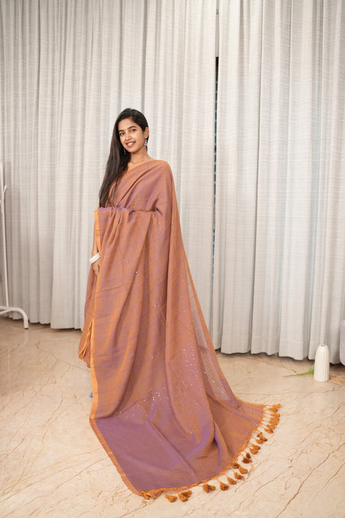 A woman modeling a sheer, lightweight saree in a dusty rose-brown color with a contrasting purple border and tassel trim, set against a neutral curtain backdrop.
